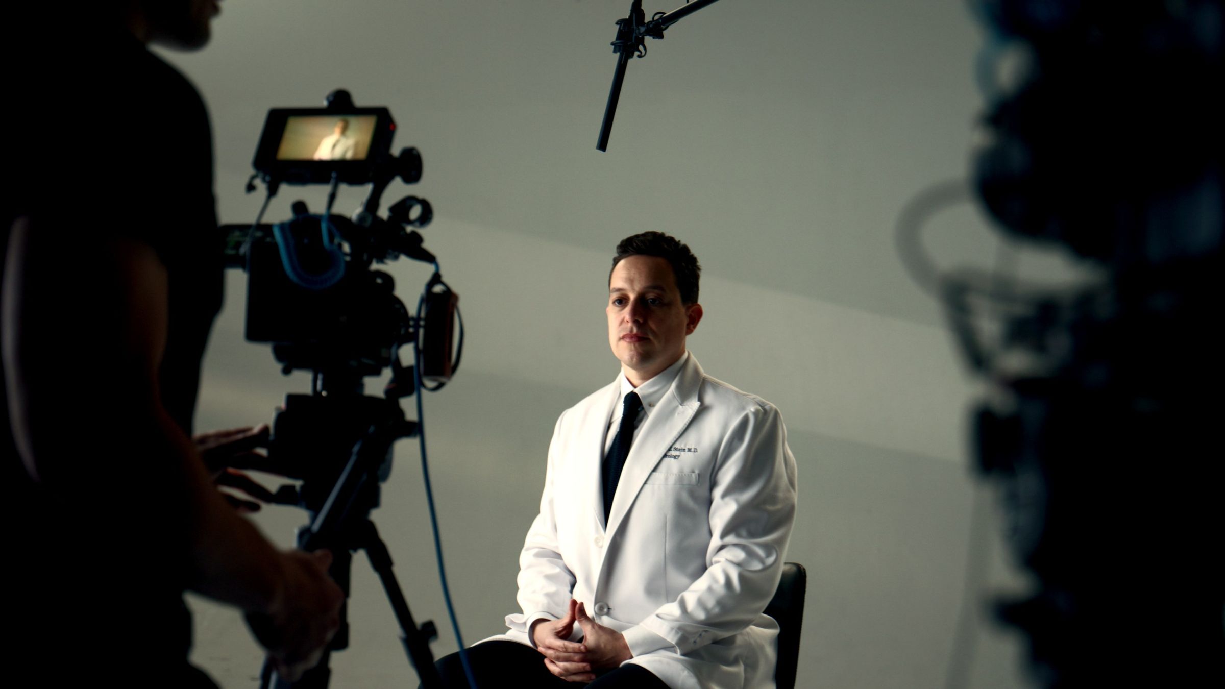 Dr. Marshall Stein in a white coat during an interview, with a cinema camera in the foreground