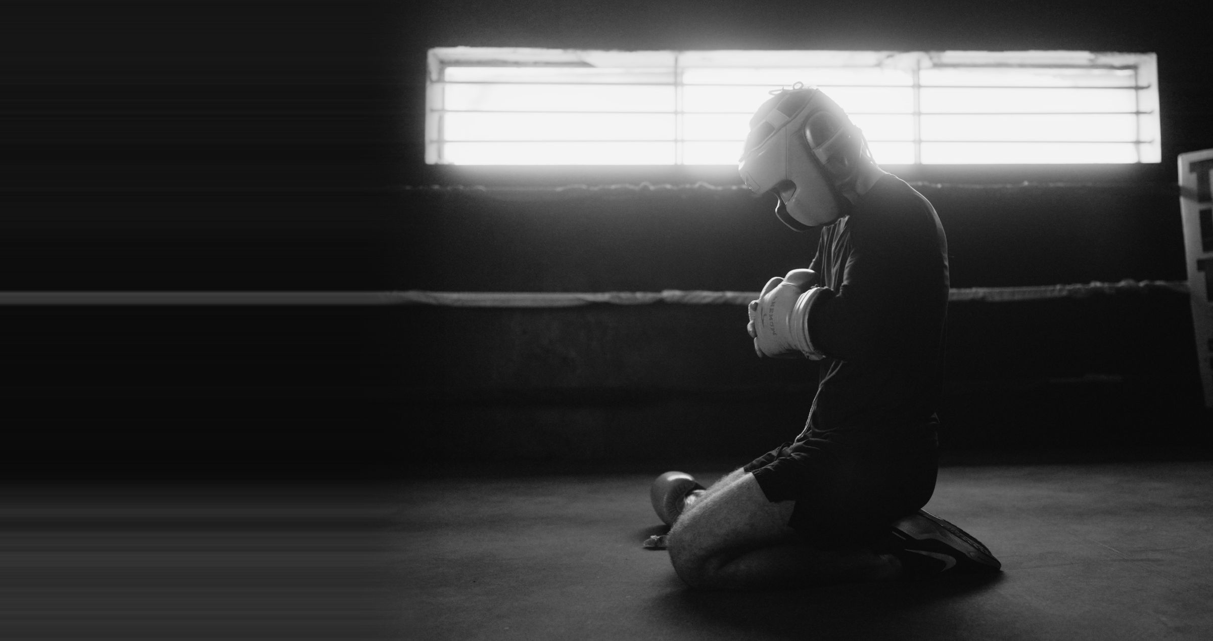 A fighter kneels in a boxing ring, lit by a window behind him