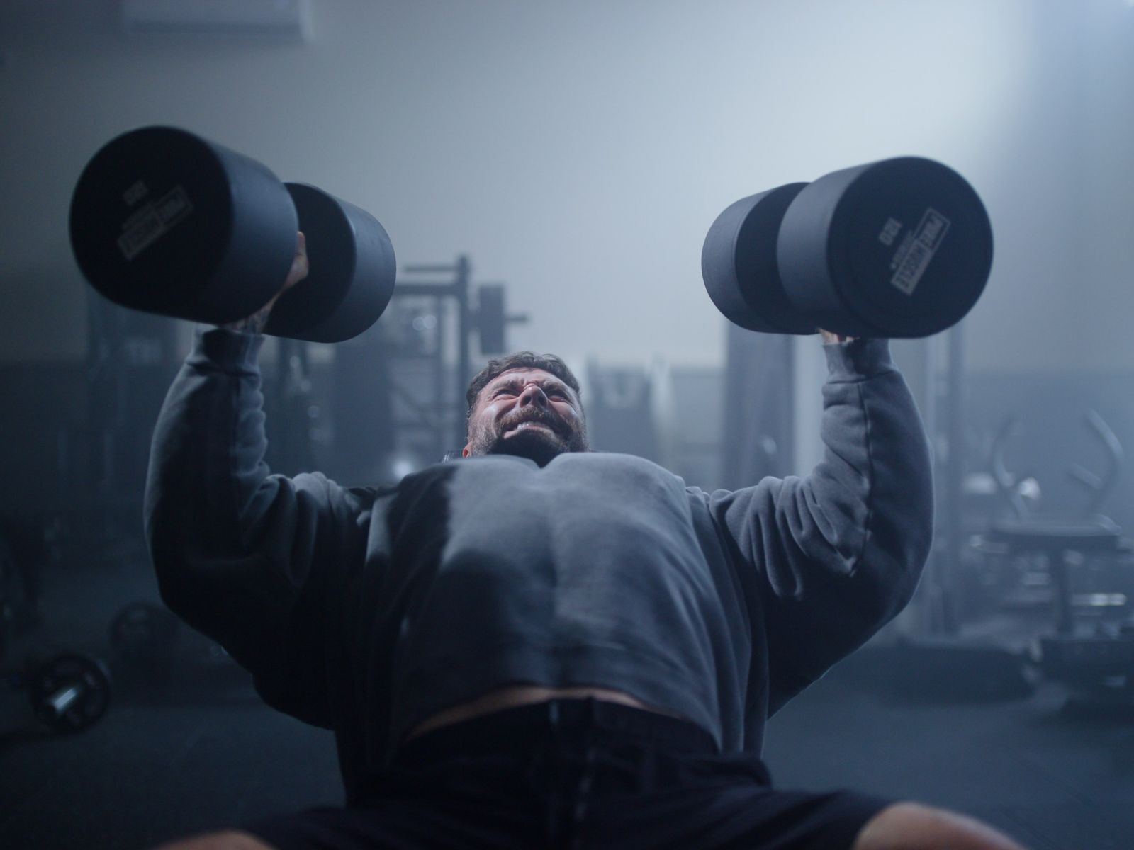 An athlete presses dumbbells in a dark gym